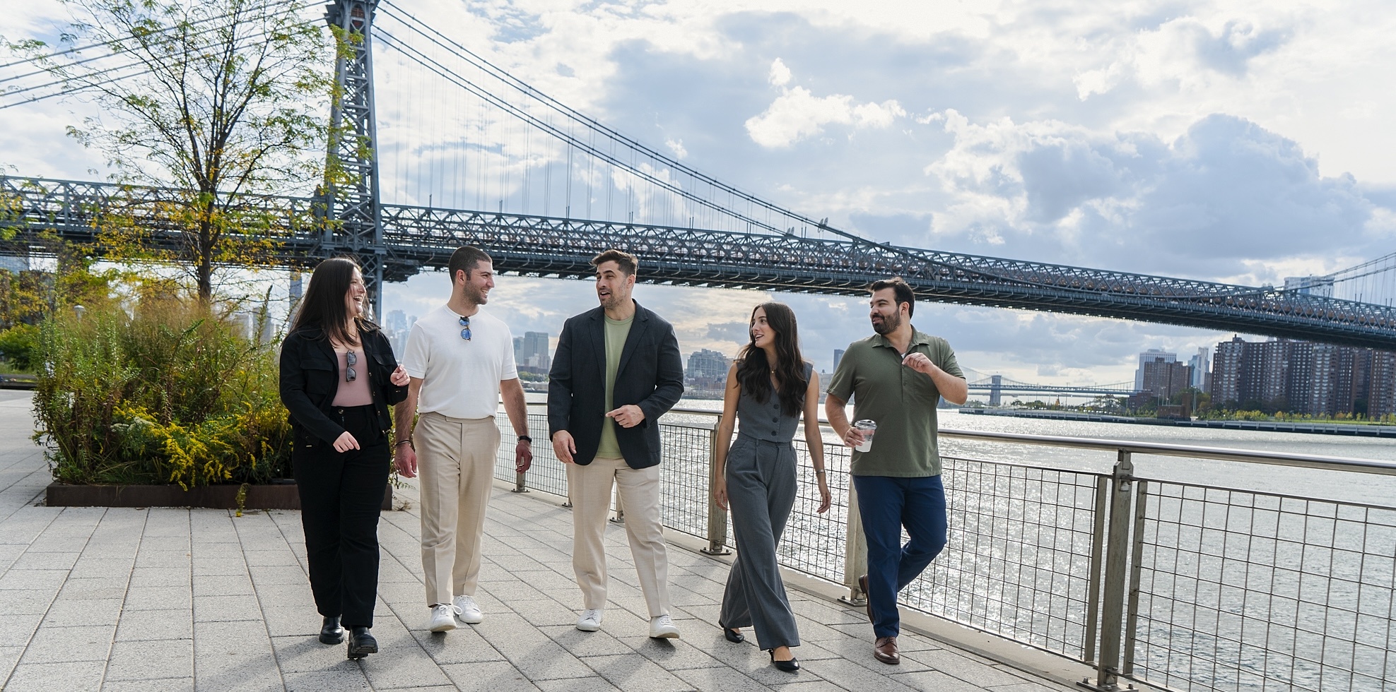 Five people walking and talking along a riverside path with a large suspension bridge in the background.