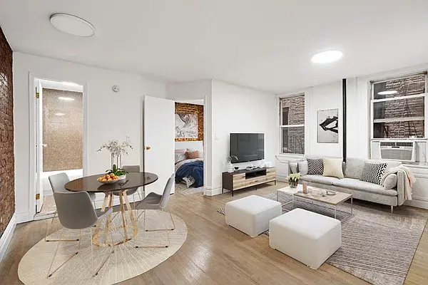Bright modern living room with a light gray sofa, white ottomans, a TV on a wooden stand, and a round dining table with chairs on a circular rug near an exposed brick wall.