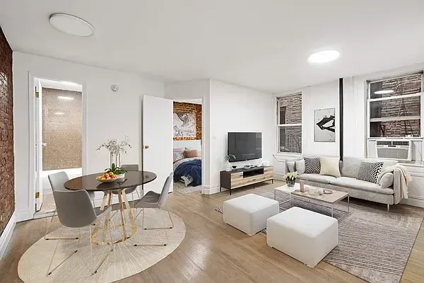 Bright modern living room with a light gray sofa, white ottomans, a TV on a wooden stand, and a round dining table with chairs on a circular rug near an exposed brick wall.