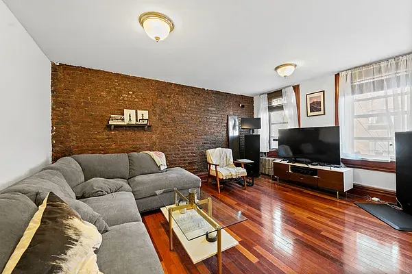 Living room with a gray sectional sofa, glass coffee table, wooden floor, brick accent wall, and TV on a modern stand.