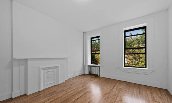 Empty room with white walls, hardwood floors, two large windows with black frames, and a white decorative fireplace.