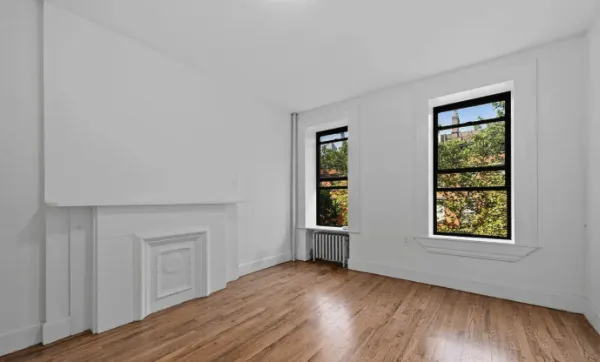 Empty room with white walls, hardwood floors, two large windows with black frames, and a white decorative fireplace.