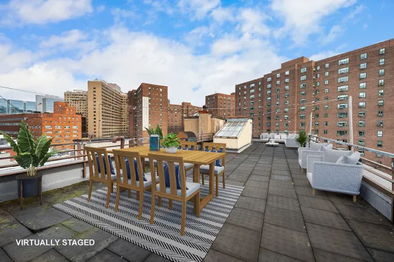 Rooftop patio with wooden dining table and chairs on a striped rug, surrounded by potted plants and gray outdoor sofas against a city skyline backdrop.
