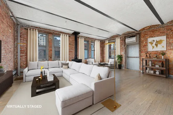 Spacious living room with exposed brick walls, large windows with curtains, a white sectional sofa, dark wooden coffee table, floor lamp, and hardwood floors.