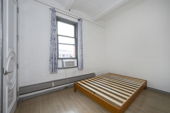 Empty bedroom with wooden slat bed frame, white walls, a window with floral curtains, and an air conditioner installed below the window.