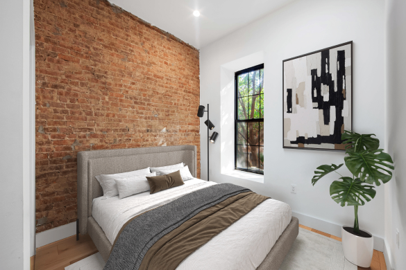 Modern bedroom with exposed brick wall, gray upholstered bed, white and brown bedding, abstract wall art, and a potted green plant near a window.