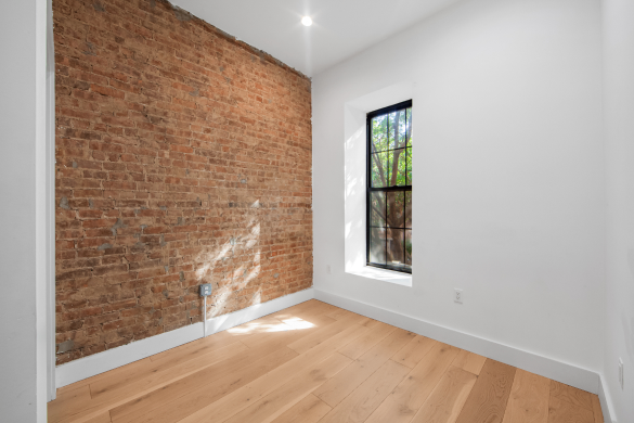 Empty room with a wooden floor, white walls, one exposed brick wall, and a window letting in natural light.