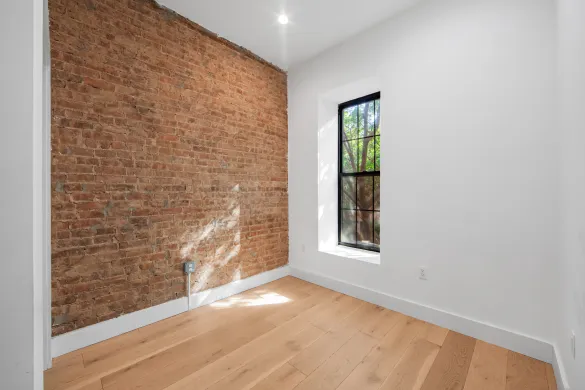 Empty room with a wooden floor, white walls, one exposed brick wall, and a window letting in natural light.