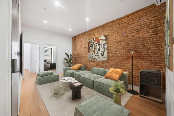 Modern living room with green sectional sofa, orange pillows, abstract wall art on exposed brick wall, and wooden floor.