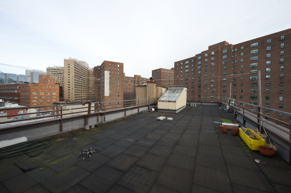 View of a rooftop with railings, skylight windows, and planters, surrounded by multiple tall brick apartment buildings under an overcast sky.