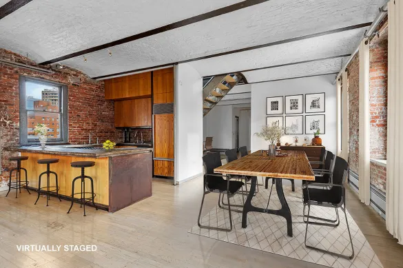 Bright loft-style kitchen and dining area with exposed brick walls, wooden kitchen island with stools, and a wooden dining table with six black chairs on a rug.