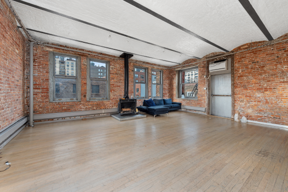Loft-style room with exposed brick walls, four windows, a black wood stove, a blue sectional sofa, and wooden floors.