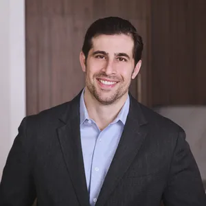 Smiling man wearing a dark blazer and white shirt standing outdoors with trees and a brick building in the background.