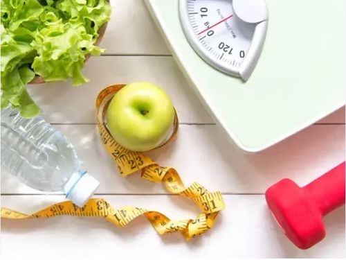 A green apple with a measuring tape around it next to a scale, hand weight, and water bottle.