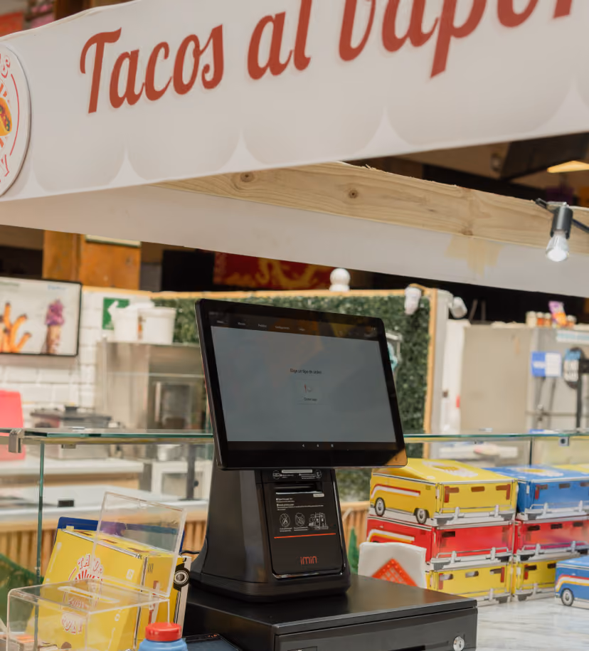 Sistema de punto de venta con pantalla táctil sobre un mostrador, debajo de un letrero que dice “Tacos al vapor”, con cajas de comida coloridas apiladas cerca en un restaurante de tacos.