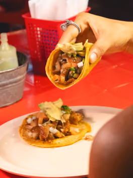 Hand holding a taco with meat and onions above a plate with another taco on a red table.