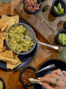 A table with guacamole, tortilla chips, salsa, lime wedges, cucumber slices, and a hand holding a chip with dip.