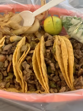 Three tacos with chopped grilled beef, onion, and a lime wedge served with onion and cilantro in a plastic container.