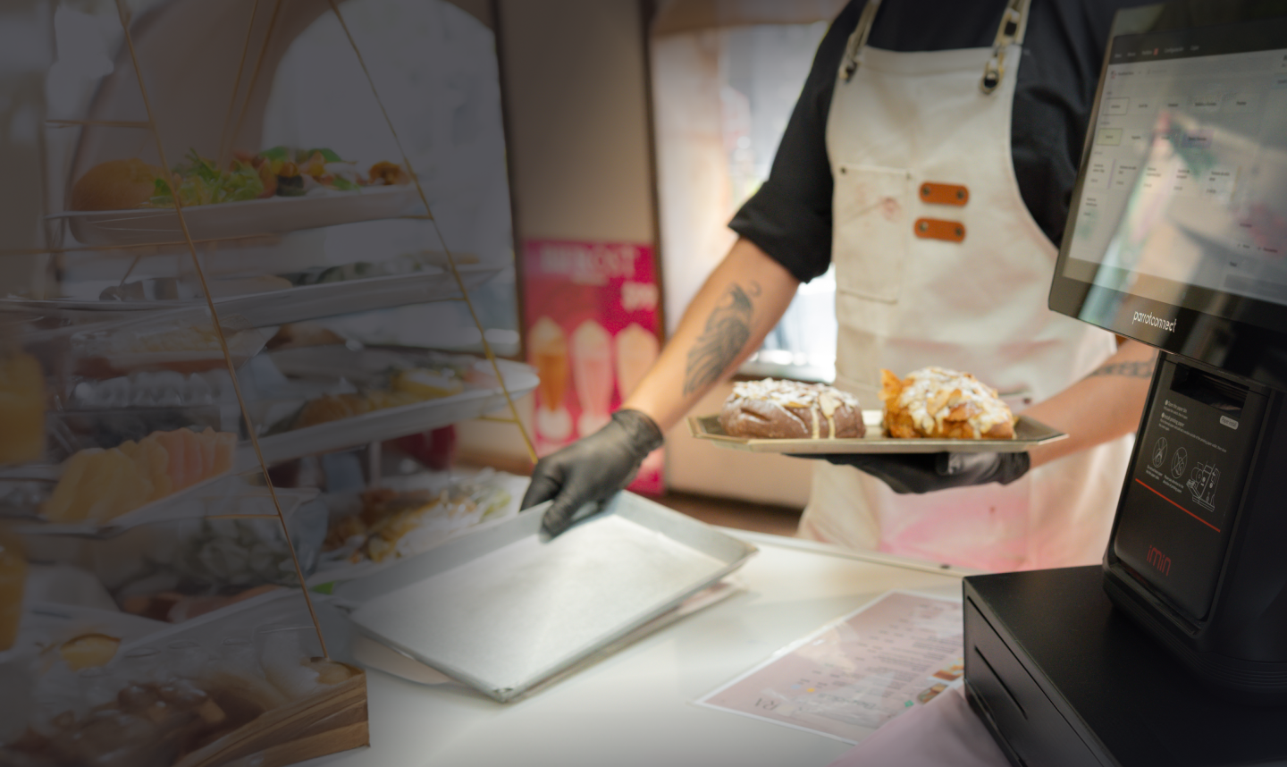 Person wearing a white apron and black gloves holding a tray with pastries behind a bakery counter near a cash register.