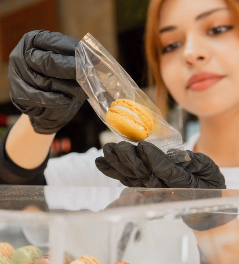 Person wearing black gloves placing a yellow macaron into a clear plastic bag.