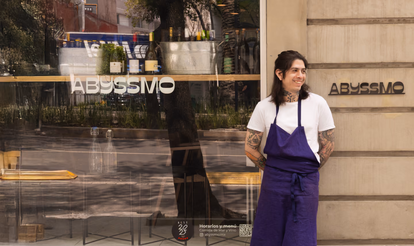 Tattooed chef in white shirt and purple apron stands smiling outside Abysmmo restaurant.