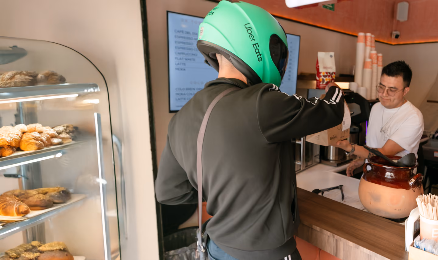Delivery person with Uber Eats helmet picks up a food order from a cafe counter employee.