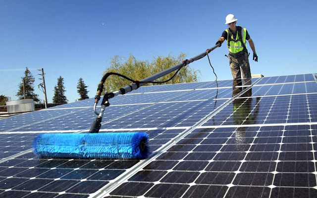 Worker in hi-vis vest and hard hat brushing rooftop solar panels with a long blue brush