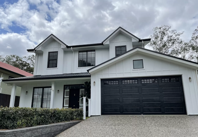 Two-storey white weatherboard home with black double garage door and pebbled driveway