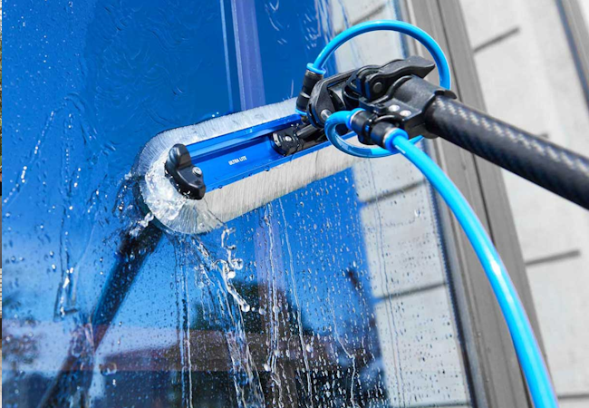 Water-fed pole brush washing a tall blue glass pane, water streaming down