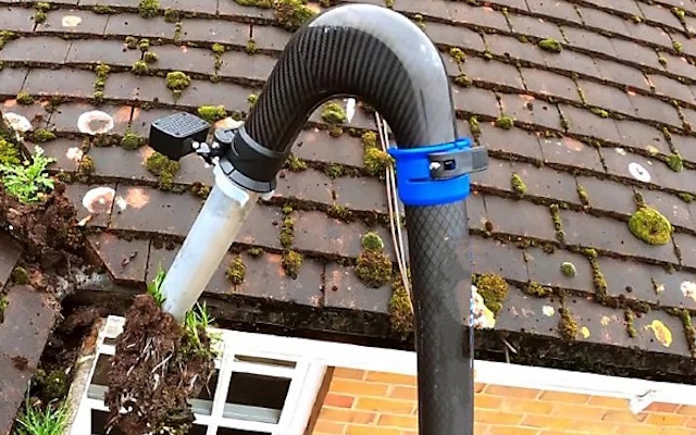 Gutter vacuum hose arched over a mossy tiled roof above a leaf-filled gutter
