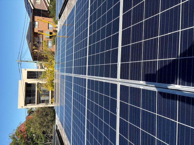 Looking along a clean solar panel array towards apartment buildings under blue sky