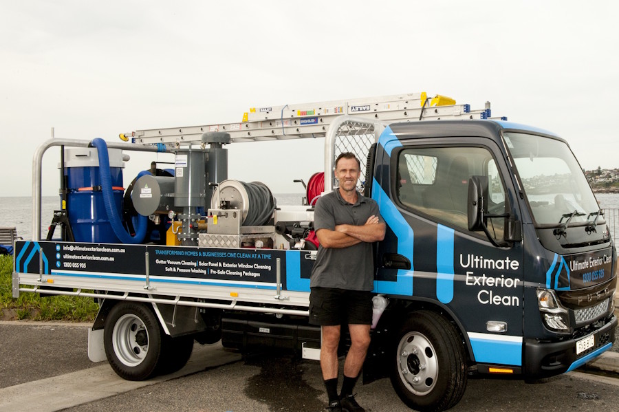 Man with crossed arms standing beside a blue and black Ultimate Exterior Clean service truck equipped with ladders and cleaning equipment, parked near a waterfront.