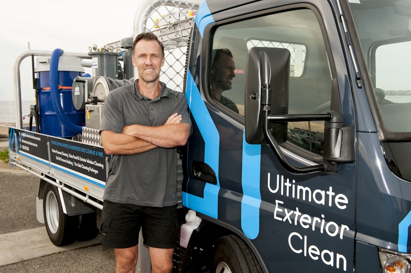 Man wearing a gray shirt and black shorts standing with arms crossed next to a blue and black truck with 'Ultimate Exterior Clean' written on the door and cleaning equipment loaded in the truck bed.