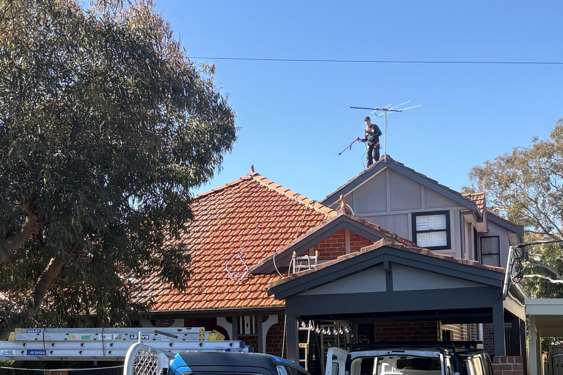 Ultimate Exterior Clean technician standing on the ridge of a terracotta tile roof in Sydney, with branded service truck and van parked below.