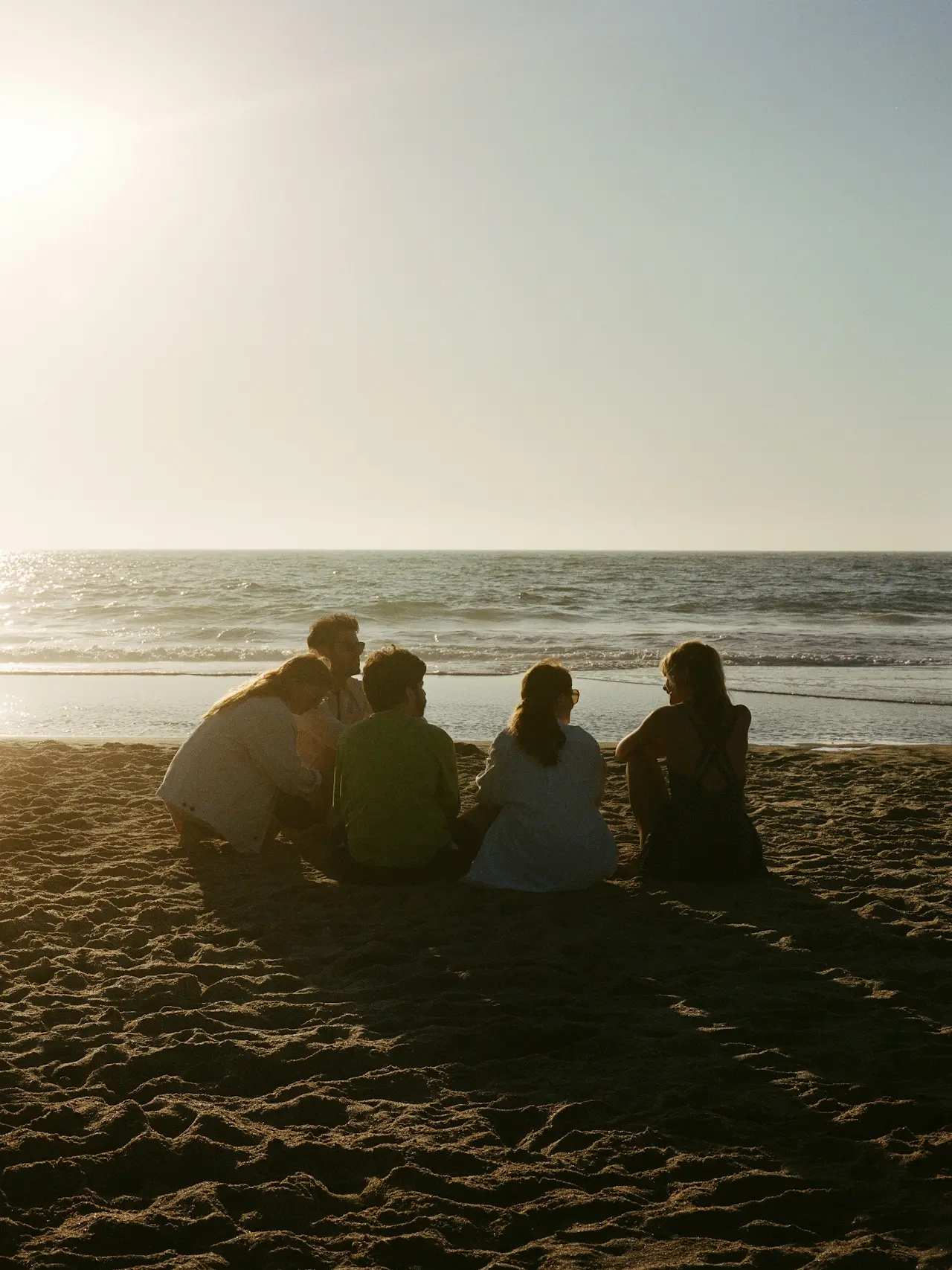 A group of people sitting on a sandy beach at sunset, facing the calm ocean. The warm, golden sunlight creates a peaceful, relaxed atmosphere.