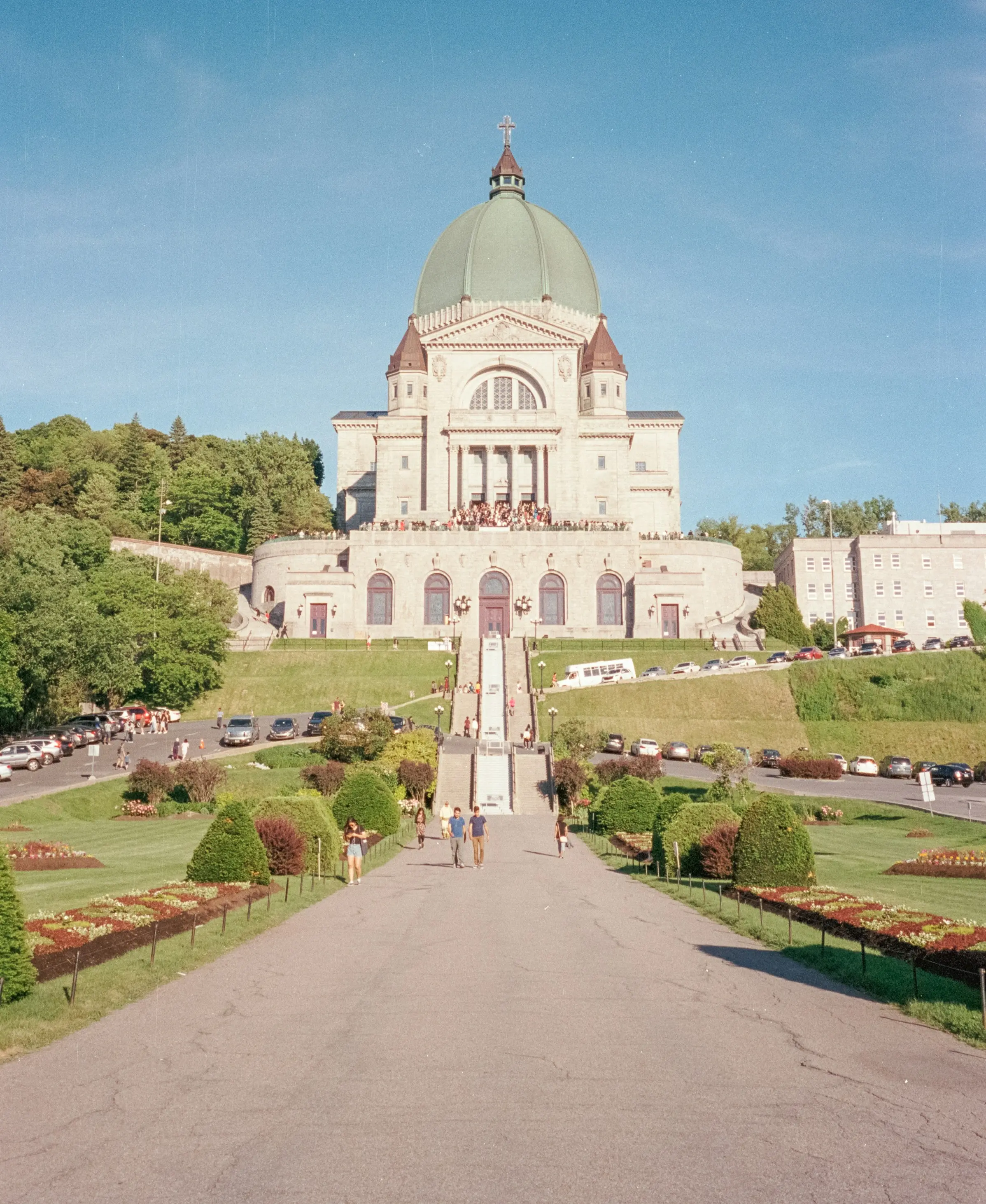 Saint Joseph's Oratory of Mount Royal, surrounded by lush gardens and trees. People walk up the steps, with clear blue skies above.