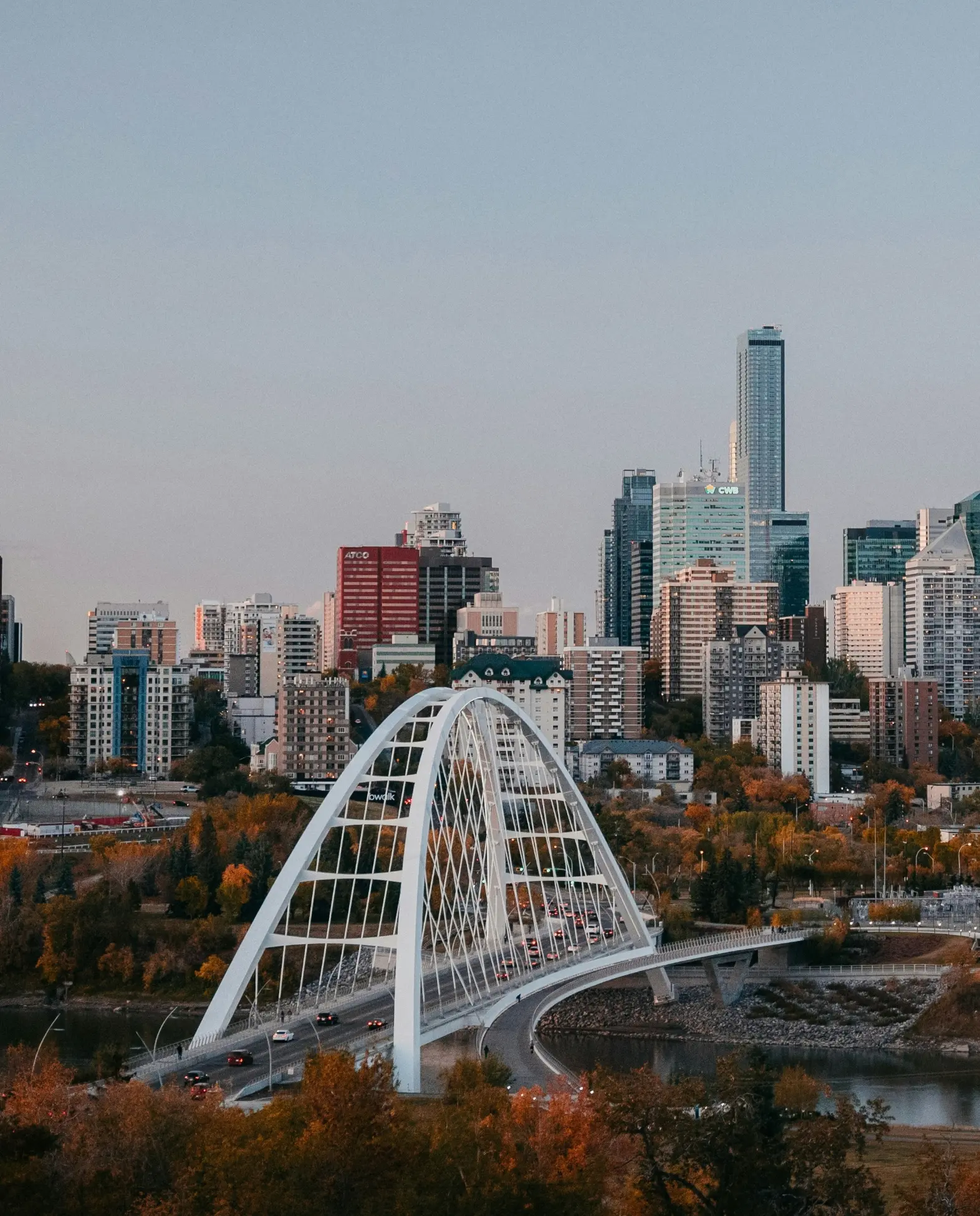 The Walterdale Bridge extends over a river, leading to a city skyline with tall skyscrapers. Autumn trees add warm colors, creating a serene urban scene.