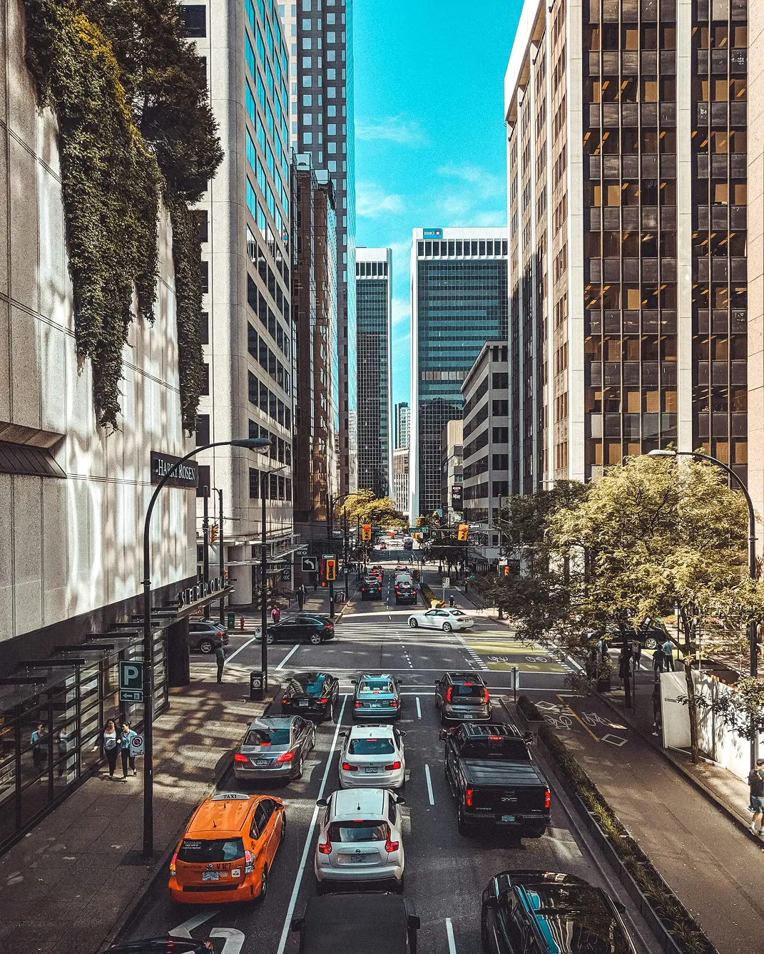 Downtown Vancouver bustling with heavy traffic, flanked by tall buildings under a clear blue sky. Trees and pedestrians add liveliness to the urban scene.