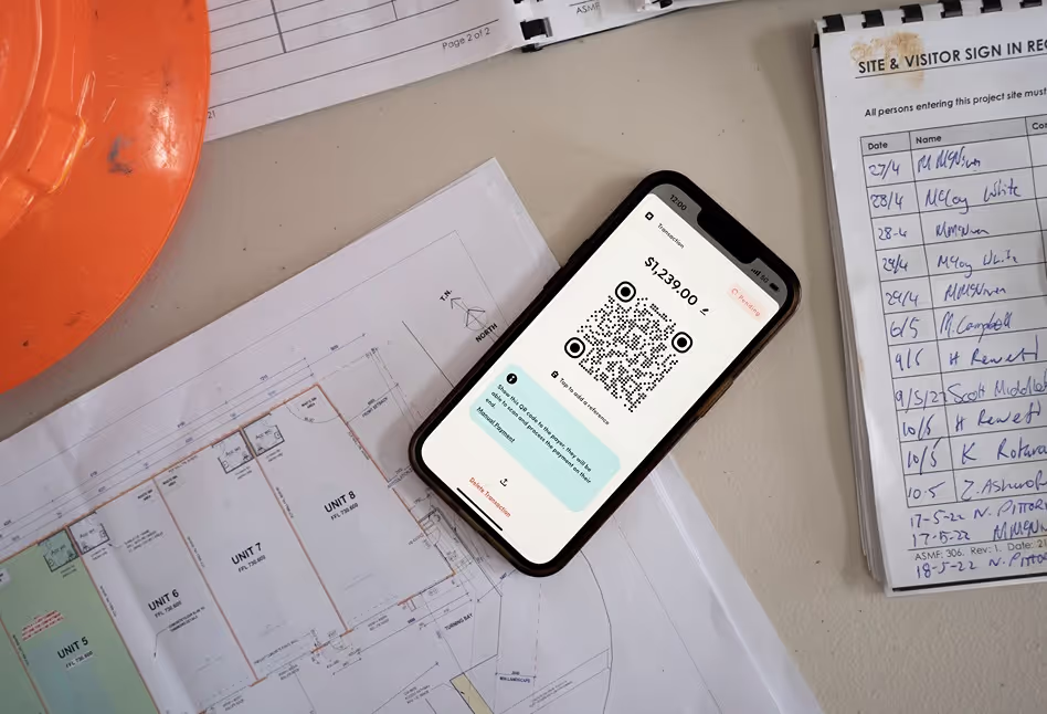 A smartphone on a desk displaying a QR code for payment, with construction plans and a visitor sign-in sheet in the background, illustrating a payment setup at a worksite.