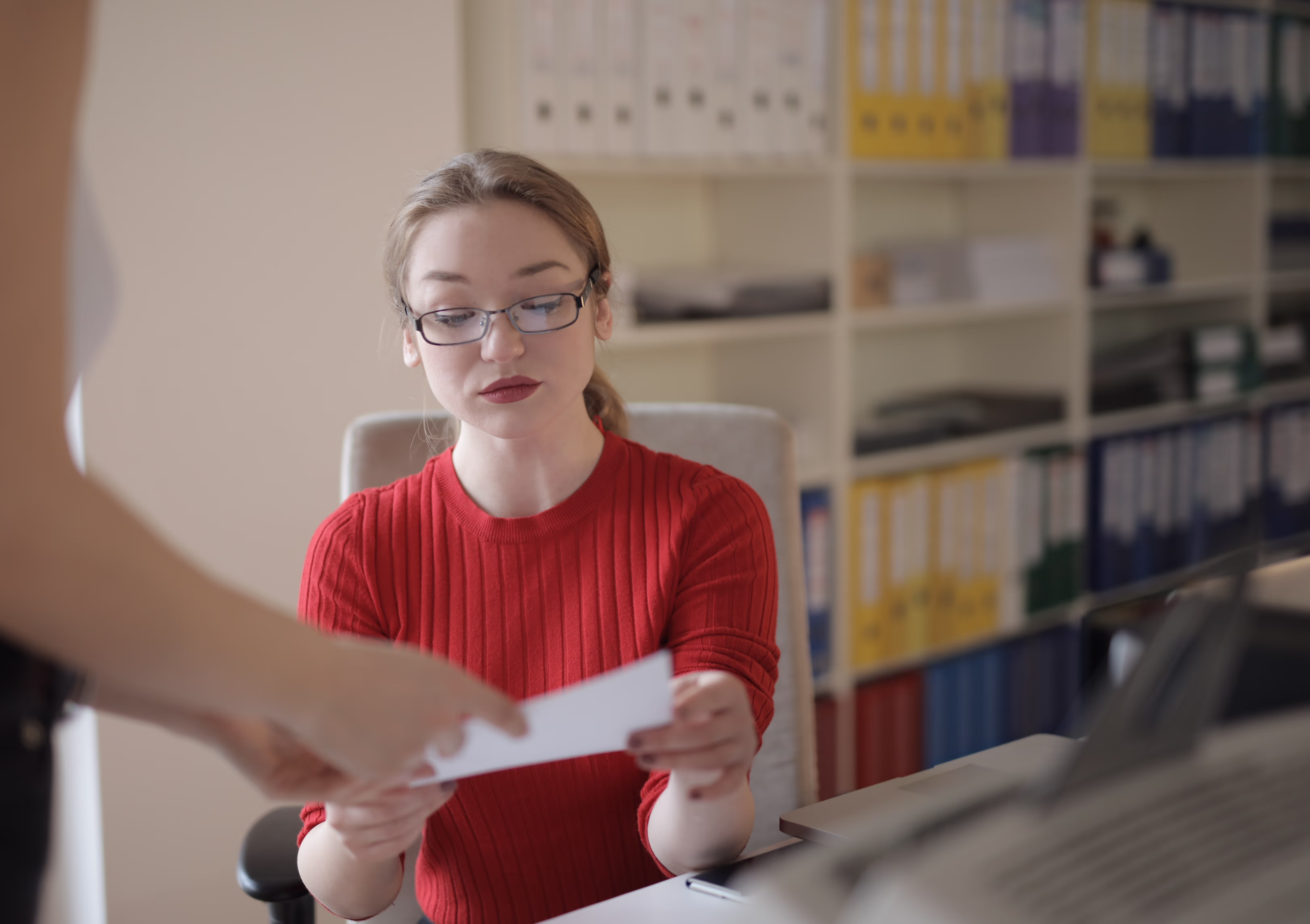 A woman in a red sweater reviews a document handed to her by another person, illustrating the ease of managing invoicing tasks in an office setting.