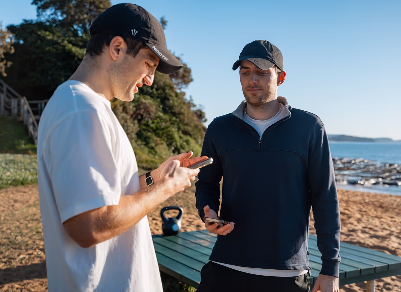 Two men in casual attire engage with 'Tap to Pay' technology on a smartphone at a sunny beachside location.