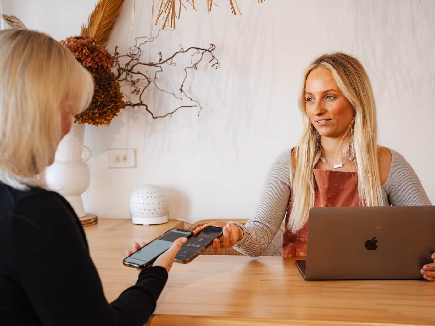Small business owner with a laptop shows the Pebl app on her phone to a customer, illustrating a modern payment method in a cozy shop setting.