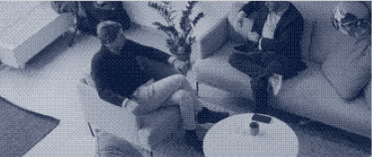 Zwei Personen sitzen auf Sesseln in einem Wohnzimmer mit Tisch und Blumenvase.