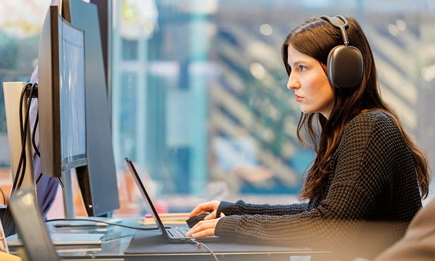 Woman wearing headphones focuses on laptop in front of multiple monitors in modern office.