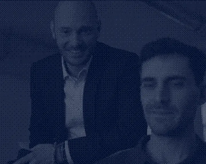 Two smiling men, one sitting in the foreground and the other in the background, both wearing formal clothes.