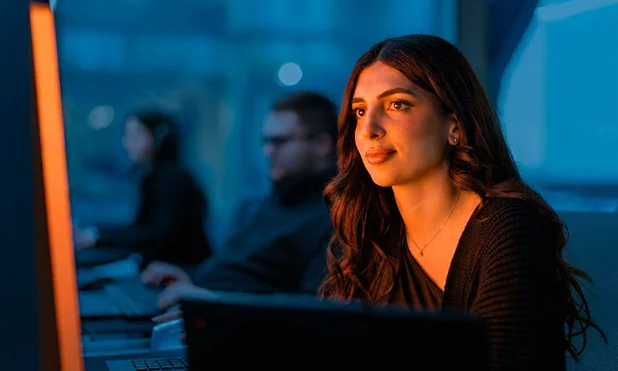 In the evening, young woman with long brown hair and a black sweater works concentrated on a computer in a dark room, with two other people blurry on computers in the background.