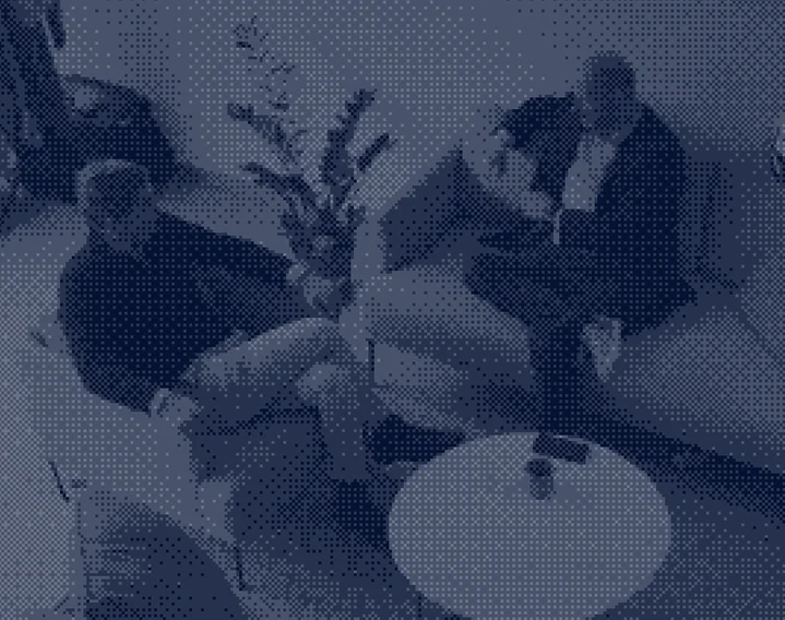 Two men sit across from each other in a modern living room on armchairs, with a small round table between them.