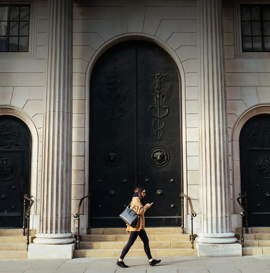 A woman walking in front of a grand building with tall columns and large black metal doors decorated with embossed symbols, carrying a bag and looking at her phone.