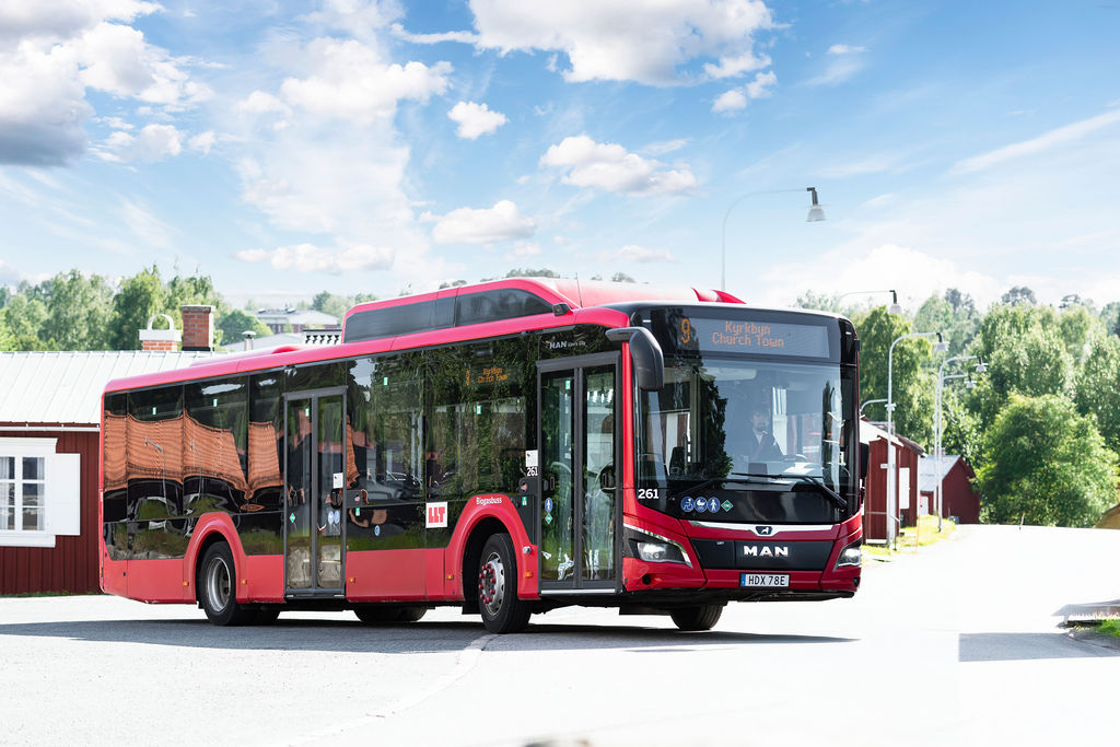 Bild på en röd LLT buss av märket MAN. I bakgrunden syns en röd kyrkstuga och blå himmel med vital moln.