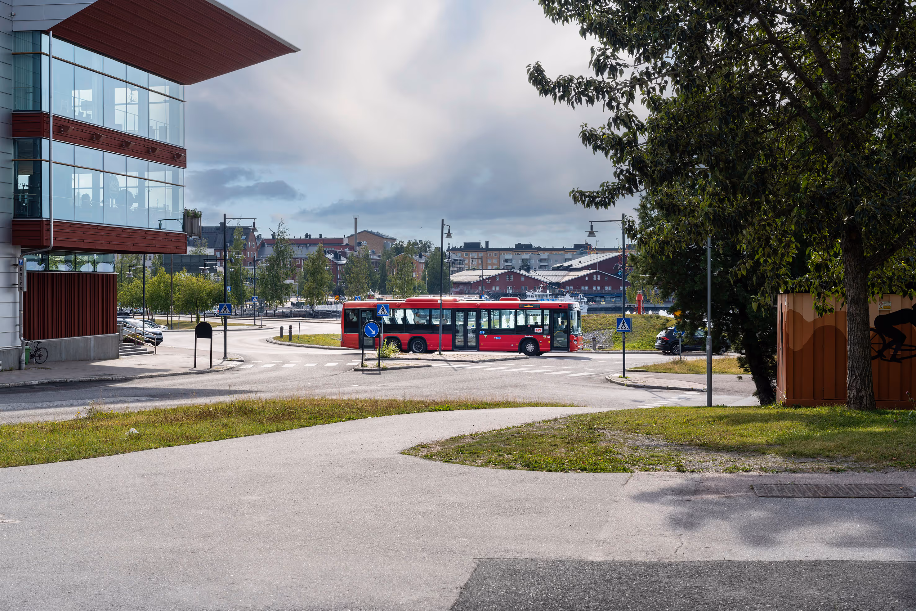 Röd stadstrafikbuss på en gata med korsning i en stadsområde, omgiven av moderna byggnader och träd.